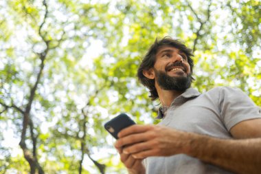 Gündüz vakti arka planda yeşil park olan akıllı telefon kullanan genç bir adamın görüntüsü. Cep telefonu, teknoloji, şehir konsepti. Yüksek kalite fotoğraf