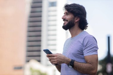 Gündüz vakti akıllı telefon kullanan ve arka planda şehir manzaralı genç bir adamın görüntüsü. Cep telefonu, teknoloji, şehir konsepti. Yüksek kalite fotoğraf