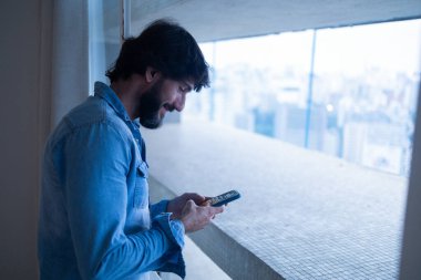 Gece vakti, arka planda şehir manzarası olan balkonunda akıllı telefon kullanan genç bir adamın yan görüntüsü. Cep telefonu, teknoloji, konsept. Telefonda mesajlaşıyoruz. Yüksek kalite fotoğraf