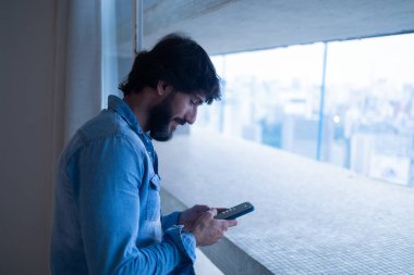 Gece vakti, arka planda şehir manzarası olan balkonunda akıllı telefon kullanan genç bir adamın yan görüntüsü. Cep telefonu, teknoloji, konsept. Telefonda mesajlaşıyoruz. Yüksek kalite fotoğraf