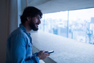 Gece vakti, arka planda şehir manzarası olan balkonunda akıllı telefon kullanan genç bir adamın yan görüntüsü. Cep telefonu, teknoloji, konsept. Telefonda mesajlaşıyoruz. Yüksek kalite fotoğraf