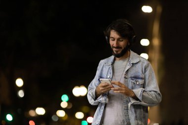 Gece vakti akıllı telefon ve kulaklık kullanan genç bir adamın ön görüntüsü. Arka planda şehir manzarası var. Yüksek kaliteli fotoğraf cep telefonu, teknoloji, konsept. Yüksek kalite fotoğraf