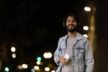 Gece vakti akıllı telefon ve kulaklık kullanan genç bir adamın ön görüntüsü. Arka planda şehir manzarası var. Yüksek kaliteli fotoğraf cep telefonu, teknoloji, konsept. Yüksek kalite fotoğraf