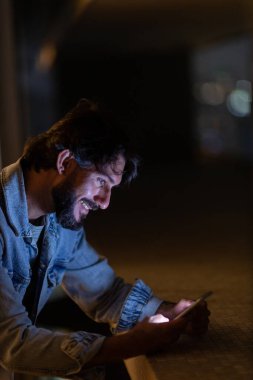Gece vakti, arka planda şehir manzarası olan balkonunda akıllı telefon kullanan genç bir adamın yan görüntüsü. Cep telefonu, teknoloji, konsept. Yüksek kalite fotoğraf