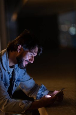 Gece vakti, arka planda şehir manzarası olan balkonunda akıllı telefon kullanan genç bir adamın yan görüntüsü. Cep telefonu, teknoloji, konsept. Yüksek kalite fotoğraf