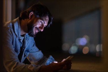 Gece vakti, arka planda şehir manzarası olan balkonunda akıllı telefon kullanan genç bir adamın yan görüntüsü. Cep telefonu, teknoloji, konsept. Yüksek kalite fotoğraf