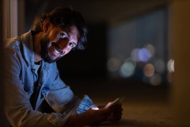 Gece vakti, arka planda şehir manzarası olan balkonunda akıllı telefon kullanan genç bir adamın yan görüntüsü. Cep telefonu, teknoloji, konsept. Yüksek kalite fotoğraf