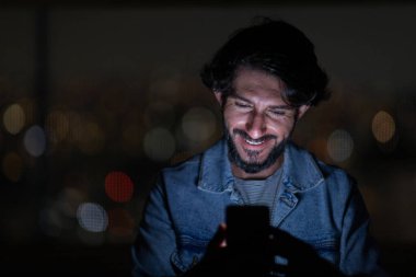 Arka planda şehir manzarası olan gece akıllı telefon kullanan genç bir adamın ön görüntüsü. Cep telefonu, teknoloji, konsept. Yüksek kalite fotoğraf