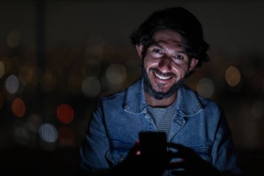 Arka planda şehir manzarası olan gece akıllı telefon kullanan genç bir adamın ön görüntüsü. Cep telefonu, teknoloji, konsept. Yüksek kalite fotoğraf