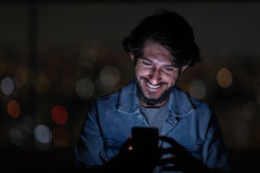 Arka planda şehir manzarası olan gece akıllı telefon kullanan genç bir adamın ön görüntüsü. Cep telefonu, teknoloji, konsept. Yüksek kalite fotoğraf