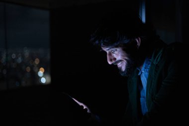 Gece vakti, arka planda şehir manzarası olan balkonunda akıllı telefon kullanan genç bir adamın yan görüntüsü. Cep telefonu, teknoloji, konsept. Yüksek kalite fotoğraf