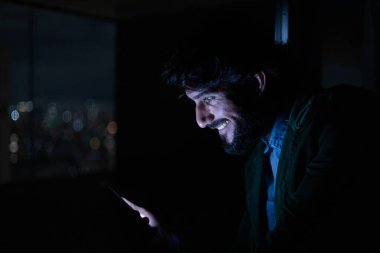 Gece vakti, arka planda şehir manzarası olan balkonunda akıllı telefon kullanan genç bir adamın yan görüntüsü. Cep telefonu, teknoloji, konsept. Yüksek kalite fotoğraf