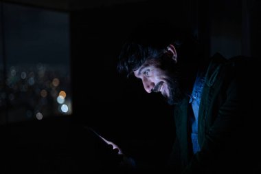 Gece vakti, arka planda şehir manzarası olan balkonunda akıllı telefon kullanan genç bir adamın yan görüntüsü. Cep telefonu, teknoloji, konsept. Yüksek kalite fotoğraf