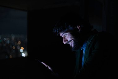 Gece vakti, arka planda şehir manzarası olan balkonunda akıllı telefon kullanan genç bir adamın yan görüntüsü. Cep telefonu, teknoloji, konsept. Yüksek kalite fotoğraf