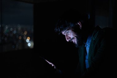 Gece vakti, arka planda şehir manzarası olan balkonunda akıllı telefon kullanan genç bir adamın yan görüntüsü. Cep telefonu, teknoloji, konsept. Yüksek kalite fotoğraf