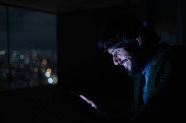 Gece vakti, arka planda şehir manzarası olan balkonunda akıllı telefon kullanan genç bir adamın yan görüntüsü. Cep telefonu, teknoloji, konsept. Yüksek kalite fotoğraf