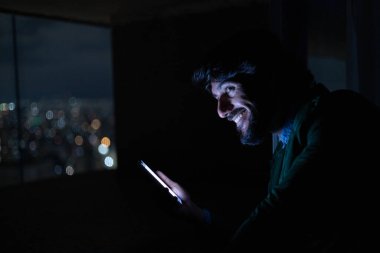 Gece vakti, arka planda şehir manzarası olan balkonunda akıllı telefon kullanan genç bir adamın yan görüntüsü. Cep telefonu, teknoloji, konsept. Yüksek kalite fotoğraf