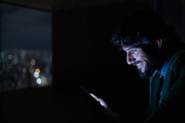 Gece vakti, arka planda şehir manzarası olan balkonunda akıllı telefon kullanan genç bir adamın yan görüntüsü. Cep telefonu, teknoloji, konsept. Yüksek kalite fotoğraf