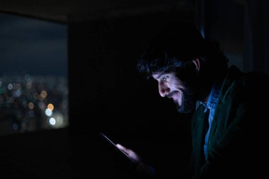 Gece vakti, arka planda şehir manzarası olan balkonunda akıllı telefon kullanan genç bir adamın yan görüntüsü. Cep telefonu, teknoloji, konsept. Yüksek kalite fotoğraf