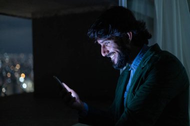 Gece vakti, arka planda şehir manzarası olan balkonunda akıllı telefon kullanan genç bir adamın yan görüntüsü. Cep telefonu, teknoloji, konsept. Yüksek kalite fotoğraf