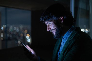 Gece vakti, arka planda şehir manzarası olan balkonunda akıllı telefon kullanan genç bir adamın yan görüntüsü. Cep telefonu, teknoloji, konsept. Yüksek kalite fotoğraf