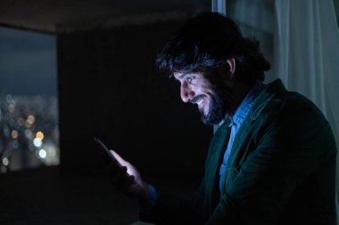 Gece vakti, arka planda şehir manzarası olan balkonunda akıllı telefon kullanan genç bir adamın yan görüntüsü. Cep telefonu, teknoloji, konsept. Yüksek kalite fotoğraf