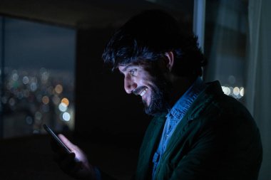 Gece vakti, arka planda şehir manzarası olan balkonunda akıllı telefon kullanan genç bir adamın yan görüntüsü. Cep telefonu, teknoloji, konsept. Yüksek kalite fotoğraf