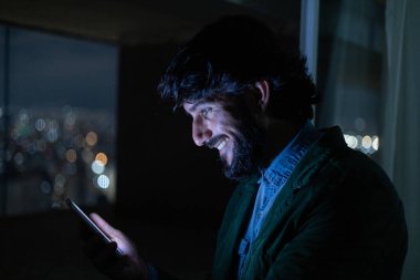 Gece vakti, arka planda şehir manzarası olan balkonunda akıllı telefon kullanan genç bir adamın yan görüntüsü. Cep telefonu, teknoloji, konsept. Yüksek kalite fotoğraf