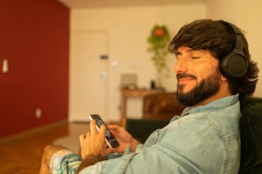 Oturma odasında kulağında kulaklıkla müzik dinleyen genç bir adamın görüntüsü. Yüksek kalite fotoğraf. Cep telefonu, teknoloji, müzik, kentsel yaşam, iyi hisset.