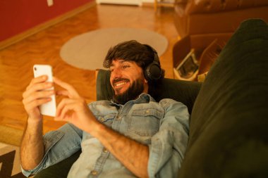 Oturma odasındaki kanepesinde uzanmış kulaklıklarla müzik dinleyen genç bir adam görüntüsü. Yüksek kalite fotoğraf. Cep telefonu, teknoloji, müzik, kentsel yaşam, iyi hisset.