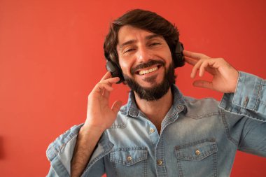 Evinde kulağında kulaklıkla müzik dinleyen genç bir adamın görüntüsü. Arka planda kırmızı boyalı duvar. Yüksek kalite fotoğraf. Cep telefonu, teknoloji, müzik, kentsel yaşam, iyi hisset.