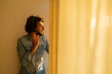 Kulağında kulaklıkla müzik dinleyen genç bir adamın yan görüntüsü pencerenin yanında arka plandaki şehir manzarasına bakıyor. Yüksek kaliteli fotoğraf cep telefonu, teknoloji, müzik, kentsel yaşam, iyi hissetme konsepti.