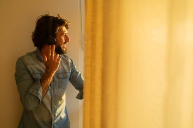 Kulağında kulaklıkla müzik dinleyen genç bir adamın yan görüntüsü pencerenin yanında arka plandaki şehir manzarasına bakıyor. Yüksek kaliteli fotoğraf cep telefonu, teknoloji, müzik, kentsel yaşam, iyi hissetme konsepti.