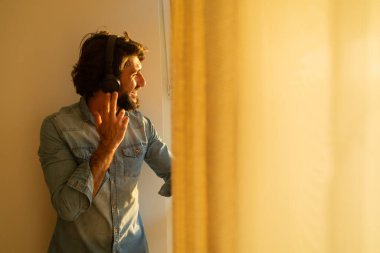 Kulağında kulaklıkla müzik dinleyen genç bir adamın yan görüntüsü pencerenin yanında arka plandaki şehir manzarasına bakıyor. Yüksek kaliteli fotoğraf cep telefonu, teknoloji, müzik, kentsel yaşam, iyi hissetme konsepti.