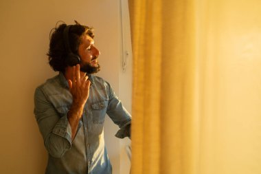 Kulağında kulaklıkla müzik dinleyen genç bir adamın yan görüntüsü pencerenin yanında arka plandaki şehir manzarasına bakıyor. Yüksek kaliteli fotoğraf cep telefonu, teknoloji, müzik, kentsel yaşam, iyi hissetme konsepti.