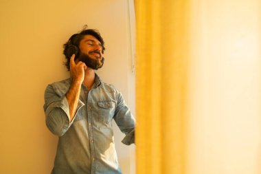 Kulağında kulaklıkla müzik dinleyen genç bir adamın yan görüntüsü pencerenin yanında arka plandaki şehir manzarasına bakıyor. Yüksek kaliteli fotoğraf cep telefonu, teknoloji, müzik, kentsel yaşam, iyi hissetme konsepti.