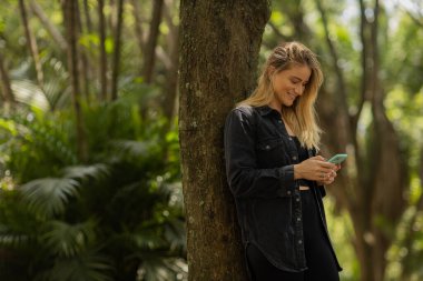 Gündüz vakti arka planda yeşil park olan akıllı telefon kullanan genç bir kadın. Yüksek kalite fotoğraf. Cep telefonu, teknoloji, şehir konsepti. Yüksek kalite fotoğraf