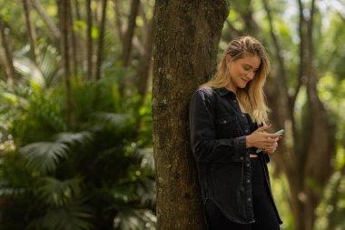 Gündüz vakti arka planda yeşil park olan akıllı telefon kullanan genç bir kadın. Yüksek kalite fotoğraf. Cep telefonu, teknoloji, şehir konsepti. Yüksek kalite fotoğraf
