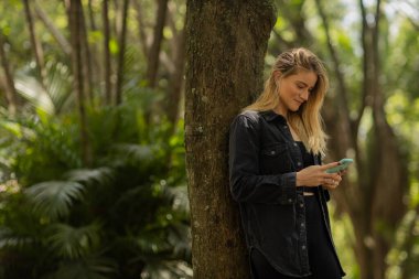 Gündüz vakti arka planda yeşil park olan akıllı telefon kullanan genç bir kadın. Yüksek kalite fotoğraf. Cep telefonu, teknoloji, şehir konsepti. Yüksek kalite fotoğraf