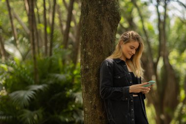 Gündüz vakti arka planda yeşil park olan akıllı telefon kullanan genç bir kadın. Yüksek kalite fotoğraf. Cep telefonu, teknoloji, şehir konsepti. Yüksek kalite fotoğraf