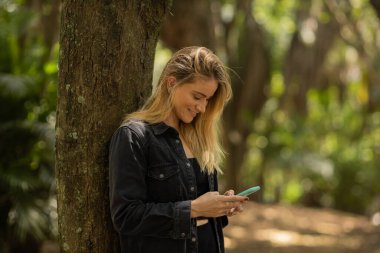 Gündüz vakti arka planda yeşil park olan akıllı telefon kullanan genç bir kadın. Yüksek kalite fotoğraf. Cep telefonu, teknoloji, şehir konsepti. Yüksek kalite fotoğraf