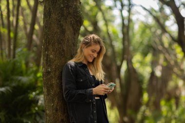 Gündüz vakti arka planda yeşil park olan akıllı telefon kullanan genç bir kadın. Yüksek kalite fotoğraf. Cep telefonu, teknoloji, şehir konsepti. Yüksek kalite fotoğraf