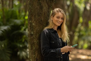 Gündüz vakti arka planda yeşil park olan akıllı telefon kullanan genç bir kadın. Yüksek kalite fotoğraf. Cep telefonu, teknoloji, şehir konsepti. Yüksek kalite fotoğraf