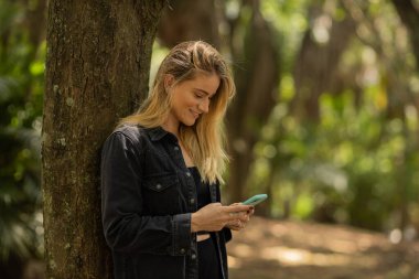 Gündüz vakti arka planda yeşil park olan akıllı telefon kullanan genç bir kadın. Yüksek kalite fotoğraf. Cep telefonu, teknoloji, şehir konsepti. Yüksek kalite fotoğraf