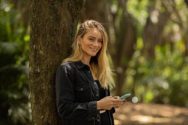 Gündüz vakti arka planda yeşil park olan akıllı telefon kullanan genç bir kadın. Yüksek kalite fotoğraf. Cep telefonu, teknoloji, şehir konsepti. Yüksek kalite fotoğraf