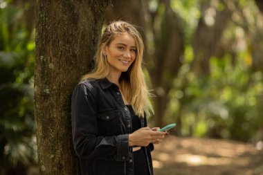 Gündüz vakti arka planda yeşil park olan akıllı telefon kullanan genç bir kadın. Yüksek kalite fotoğraf. Cep telefonu, teknoloji, şehir konsepti. Yüksek kalite fotoğraf