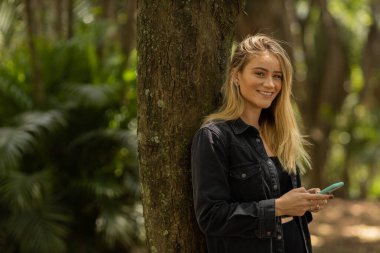 Gündüz vakti arka planda yeşil park olan akıllı telefon kullanan genç bir kadın. Yüksek kalite fotoğraf. Cep telefonu, teknoloji, şehir konsepti. Yüksek kalite fotoğraf
