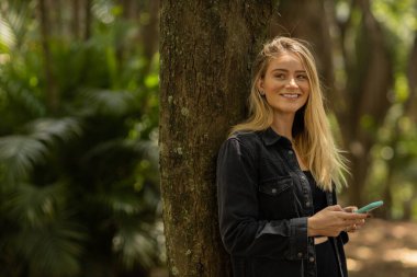 Gündüz vakti arka planda yeşil park olan akıllı telefon kullanan genç bir kadın. Yüksek kalite fotoğraf. Cep telefonu, teknoloji, şehir konsepti. Yüksek kalite fotoğraf