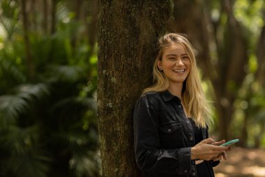 Gündüz vakti arka planda yeşil park olan akıllı telefon kullanan genç bir kadın. Yüksek kalite fotoğraf. Cep telefonu, teknoloji, şehir konsepti. Yüksek kalite fotoğraf