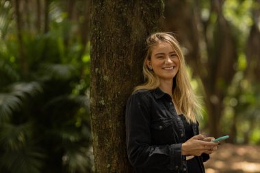 Gündüz vakti arka planda yeşil park olan akıllı telefon kullanan genç bir kadın. Yüksek kalite fotoğraf. Cep telefonu, teknoloji, şehir konsepti. Yüksek kalite fotoğraf